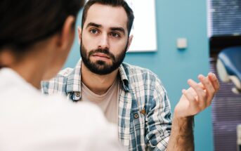 A bearded man with short hair and wearing a plaid shirt gestures with his hand while listening intently to another person. They are indoors, with a blue wall and window blinds in the background.