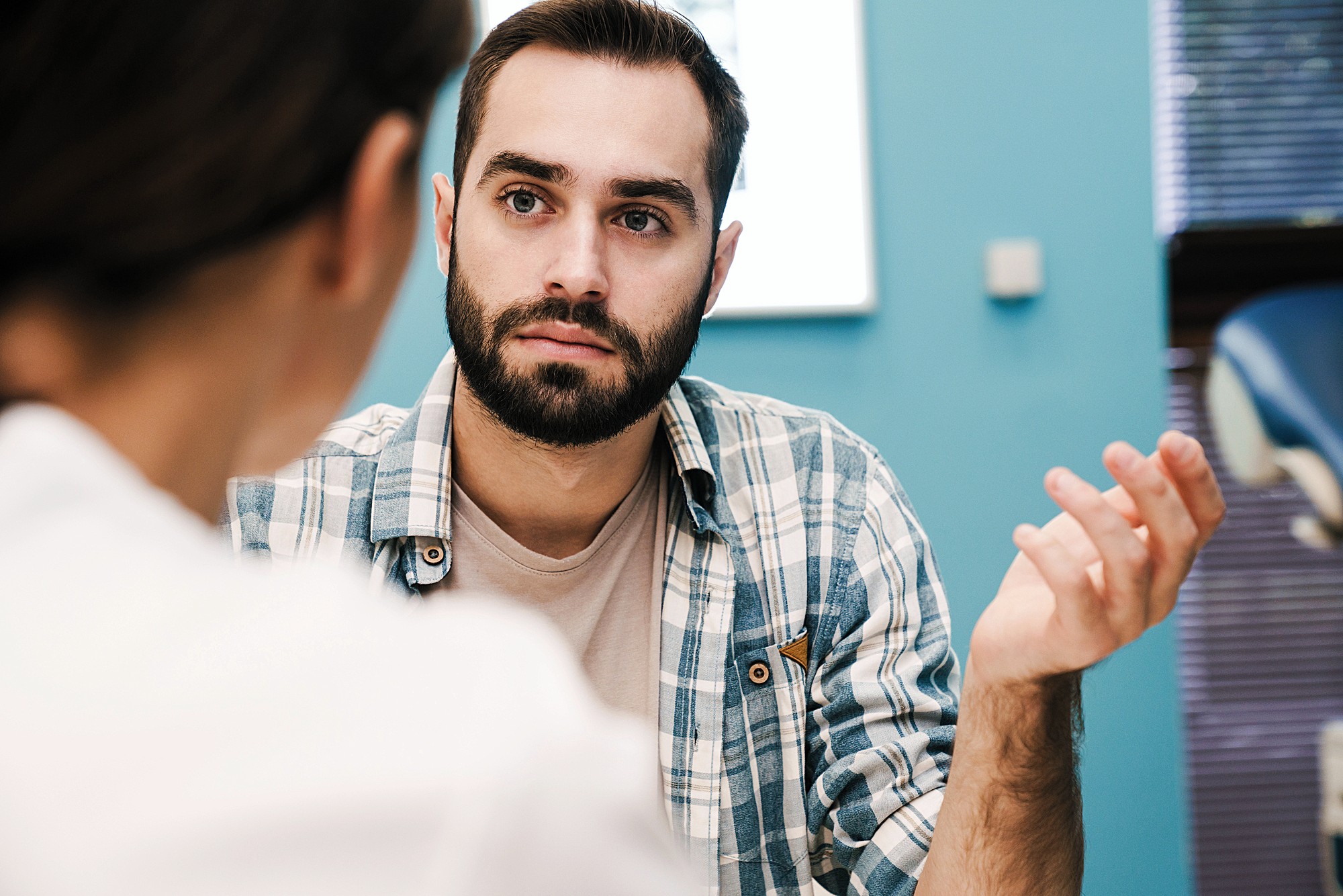 A bearded man with short hair and wearing a plaid shirt gestures with his hand while listening intently to another person. They are indoors, with a blue wall and window blinds in the background.