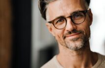 A man with gray hair and a beard, wearing glasses and a beige sweater, smiles softly at the camera. The background is blurred, emphasizing his casual and friendly expression.