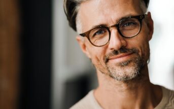 A man with gray hair and a beard, wearing glasses and a beige sweater, smiles softly at the camera. The background is blurred, emphasizing his casual and friendly expression.