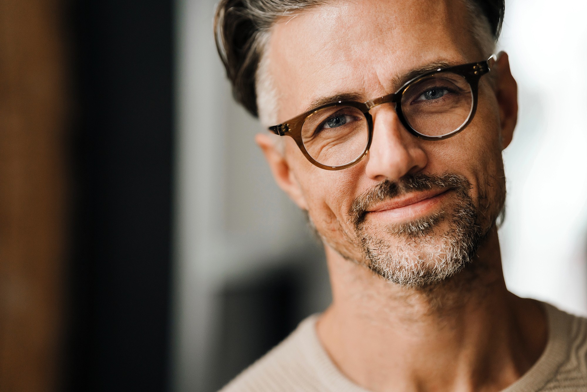 A man with gray hair and a beard, wearing glasses and a beige sweater, smiles softly at the camera. The background is blurred, emphasizing his casual and friendly expression.