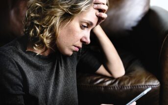 Woman with curly hair looks at a smartphone while resting her head on her hand, sitting on a brown leather couch. She appears deep in thought or concerned.