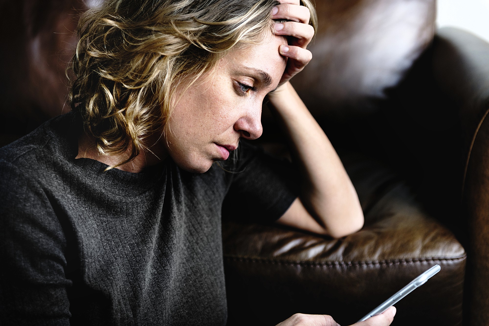 Woman with curly hair looks at a smartphone while resting her head on her hand, sitting on a brown leather couch. She appears deep in thought or concerned.
