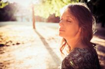 A woman with long hair stands outdoors with her eyes closed, basking in the warm sunlight. She is wearing a patterned top and appears relaxed. The sun shines through the trees, casting a gentle glow over the scene.