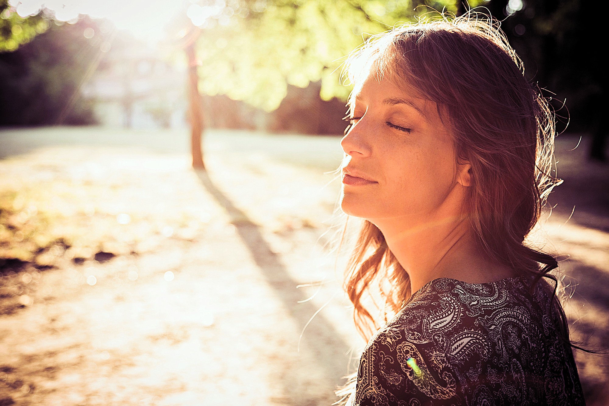 A woman with long hair stands outdoors with her eyes closed, basking in the warm sunlight. She is wearing a patterned top and appears relaxed. The sun shines through the trees, casting a gentle glow over the scene.