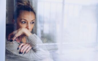 A woman with a bun is gazing thoughtfully out of a window, resting her chin on her hand. She is wearing a gray sweater and has dark nail polish. The mood is pensive, with reflections on the glass adding a layer of depth.