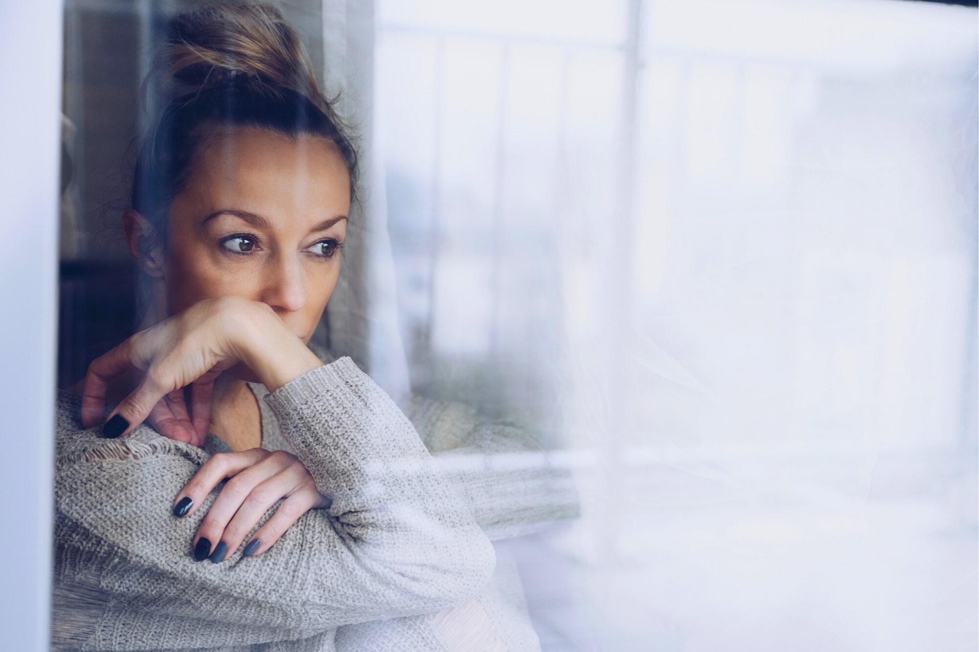 A woman with a bun is gazing thoughtfully out of a window, resting her chin on her hand. She is wearing a gray sweater and has dark nail polish. The mood is pensive, with reflections on the glass adding a layer of depth.