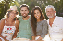 A group of four people sitting closely together outdoors. On the left, a woman in a hat and colorful top, next to a man in a green shirt. To his right, a woman in a striped top, and an older man in a white shirt. They are smiling with trees in the background.