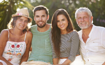 A group of four people sitting closely together outdoors. On the left, a woman in a hat and colorful top, next to a man in a green shirt. To his right, a woman in a striped top, and an older man in a white shirt. They are smiling with trees in the background.