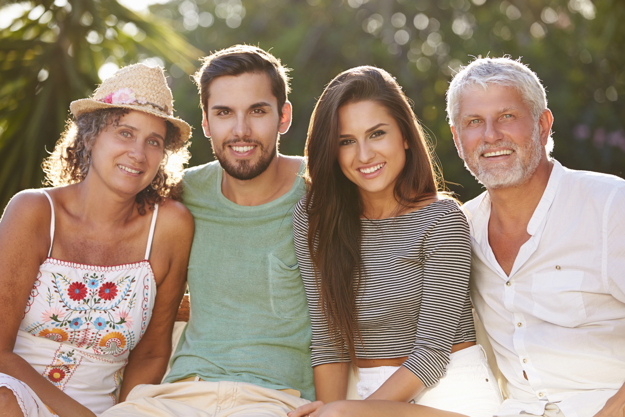 A group of four people sitting closely together outdoors. On the left, a woman in a hat and colorful top, next to a man in a green shirt. To his right, a woman in a striped top, and an older man in a white shirt. They are smiling with trees in the background.