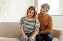 A woman in a striped shirt sits on a couch, looking down thoughtfully. A man in a brown sweater sits beside her, gently placing his hand on her knee in a comforting manner. The room is softly lit with light-colored décor.
