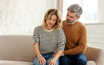 A woman in a striped shirt sits on a couch, looking down thoughtfully. A man in a brown sweater sits beside her, gently placing his hand on her knee in a comforting manner. The room is softly lit with light-colored décor.