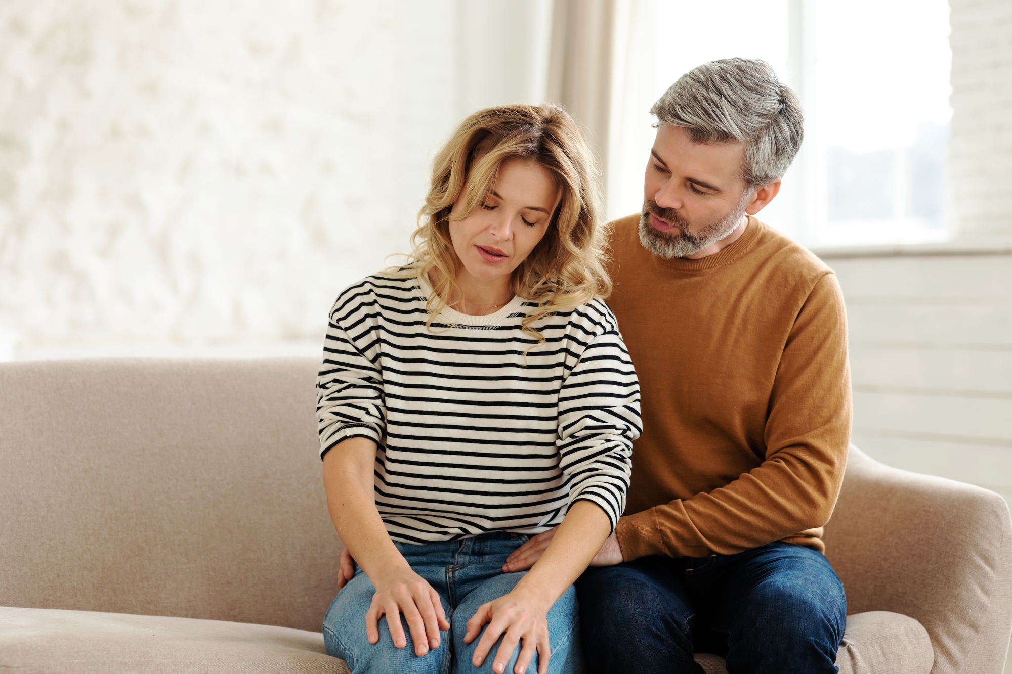 A woman in a striped shirt sits on a couch, looking down thoughtfully. A man in a brown sweater sits beside her, gently placing his hand on her knee in a comforting manner. The room is softly lit with light-colored décor.
