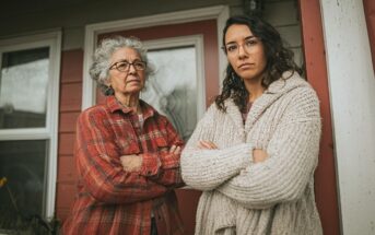 Two women with serious expressions stand outside a house with red siding and glass doors, both wearing glasses and warm clothing, with their arms crossed and looking directly at the camera.