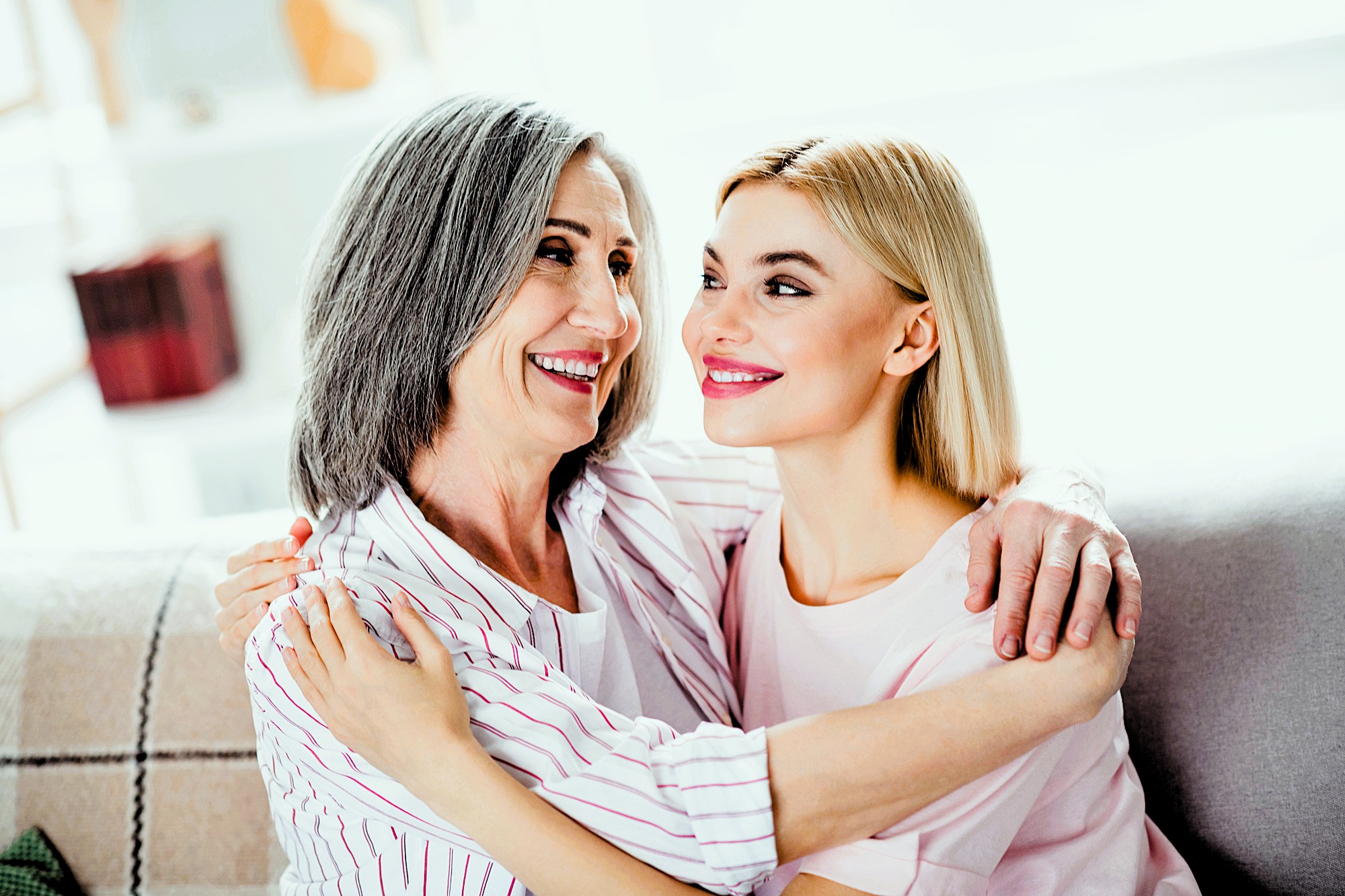 A smiling older woman with gray hair and a younger woman with blonde hair sit closely on a couch. They are embracing each other affectionately, looking at one another, with a bright and cheerful atmosphere in the room.