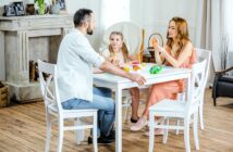 A man, a woman, and a young girl sit around a white table playing with modeling clay at home. The woman and girl are sculpting, while the man is speaking. The room has light-colored decor and a wooden floor.