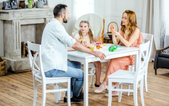 A man, a woman, and a young girl sit around a white table playing with modeling clay at home. The woman and girl are sculpting, while the man is speaking. The room has light-colored decor and a wooden floor.