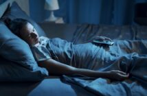 A person is sleeping in bed, covered with a dark gray blanket. They are holding a TV remote in one hand. The room is dimly lit with a lamp in the background, creating a peaceful nighttime atmosphere.