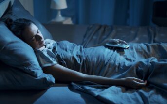 A person is sleeping in bed, covered with a dark gray blanket. They are holding a TV remote in one hand. The room is dimly lit with a lamp in the background, creating a peaceful nighttime atmosphere.