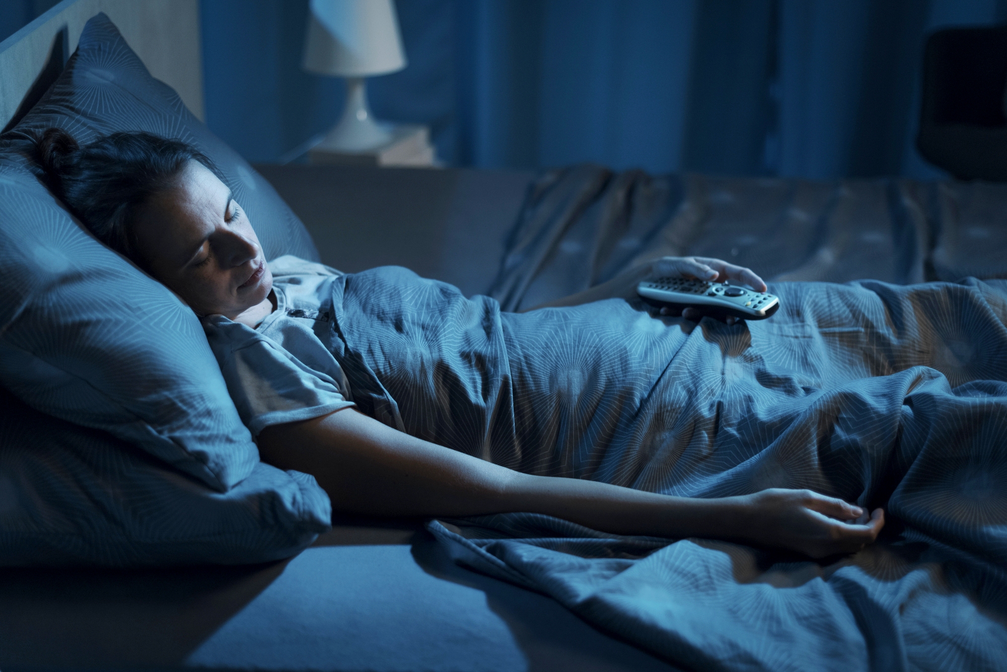 A person is sleeping in bed, covered with a dark gray blanket. They are holding a TV remote in one hand. The room is dimly lit with a lamp in the background, creating a peaceful nighttime atmosphere.