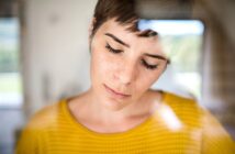 A person with short hair and closed eyes is seen through a glass window, wearing a yellow patterned top. The reflection on the glass creates a soft, dreamy effect.