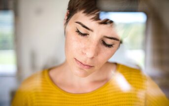 A person with short hair and closed eyes is seen through a glass window, wearing a yellow patterned top. The reflection on the glass creates a soft, dreamy effect.