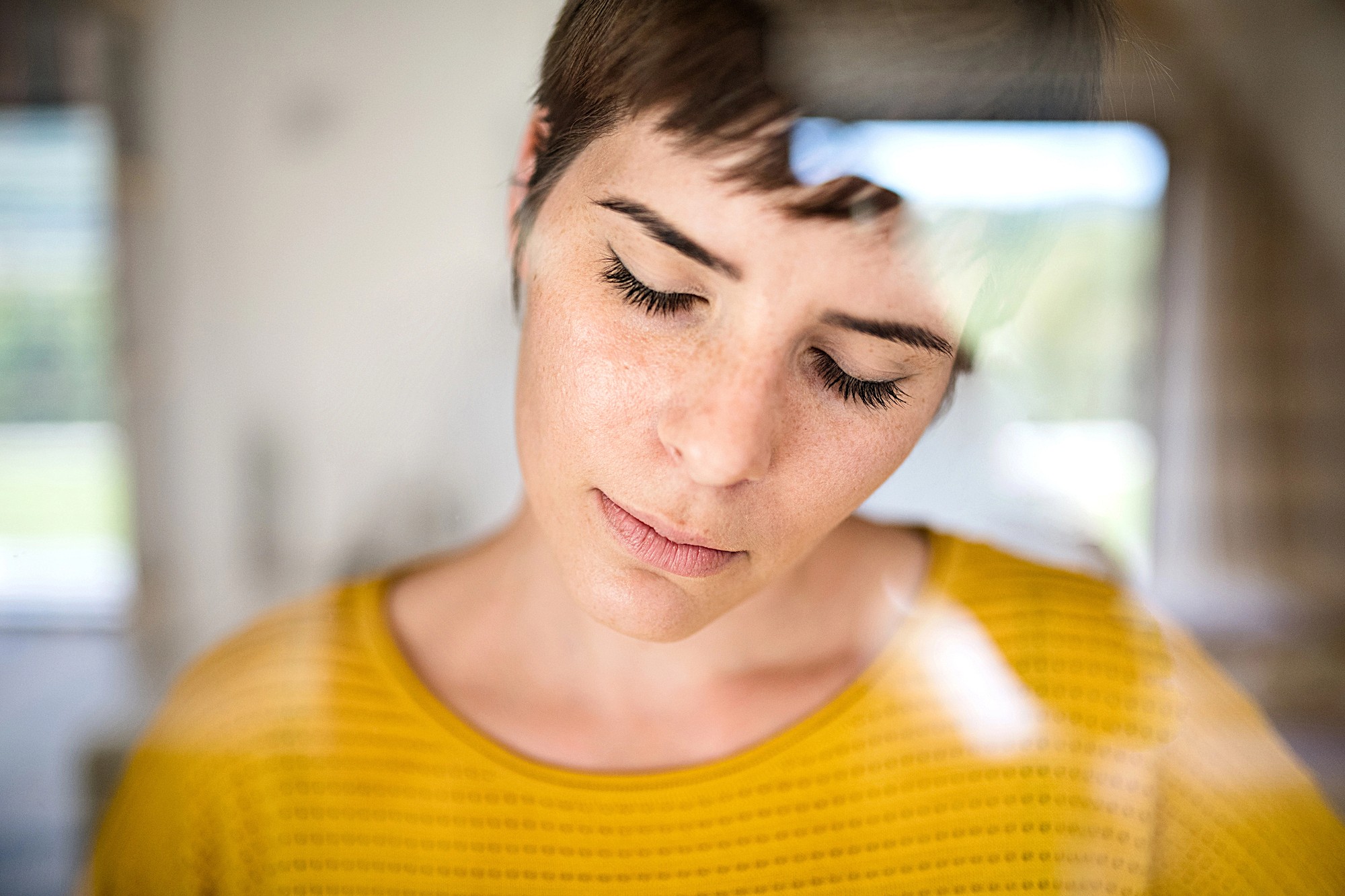 A person with short hair and closed eyes is seen through a glass window, wearing a yellow patterned top. The reflection on the glass creates a soft, dreamy effect.