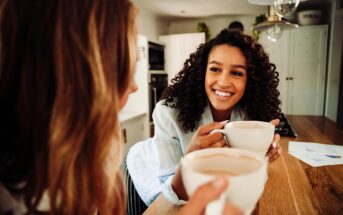 Two people are sitting in a kitchen, holding mugs and smiling at each other. One person has curly hair and is wearing a white shirt. The other has wavy hair. Papers are on the counter nearby, and the interior is warmly lit.