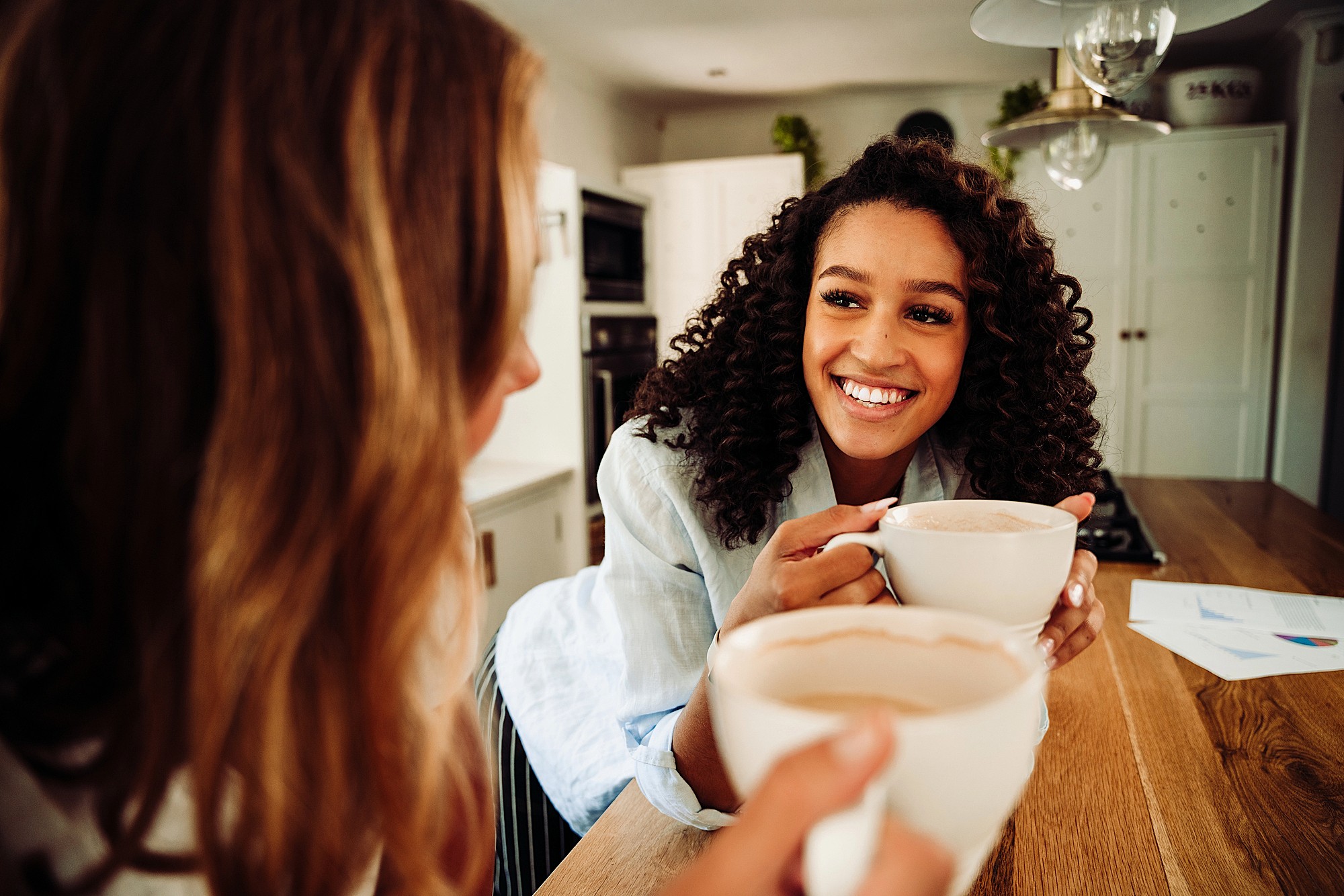 Two people are sitting in a kitchen, holding mugs and smiling at each other. One person has curly hair and is wearing a white shirt. The other has wavy hair. Papers are on the counter nearby, and the interior is warmly lit.