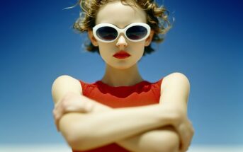 A woman wearing white sunglasses and a red sleeveless top stands with her arms crossed, looking confidently at the camera against a clear blue sky. Her curly hair is windswept and she wears bright red lipstick.