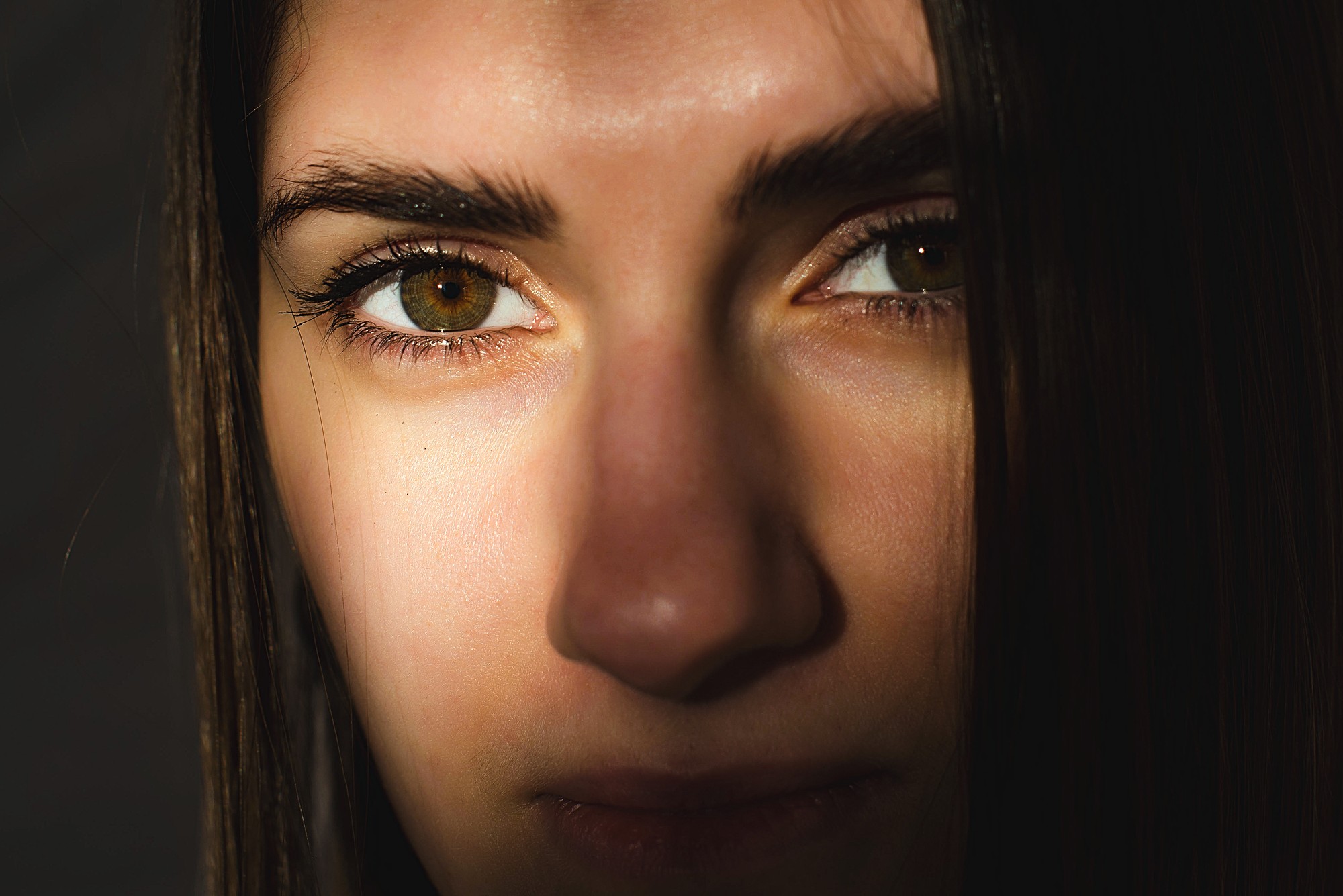 Close-up of a person's face partially in shadow. The focus is on their brown eyes and skin with soft lighting highlighting part of their forehead, nose, and lips. Hair falls on the right side of their face.