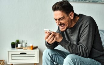 A man in a gray sweater sits on a bed, gripping a smartphone tightly. His expression is intense and frustrated. The bedside table next to him holds various small items, including a plant and a pill bottle.