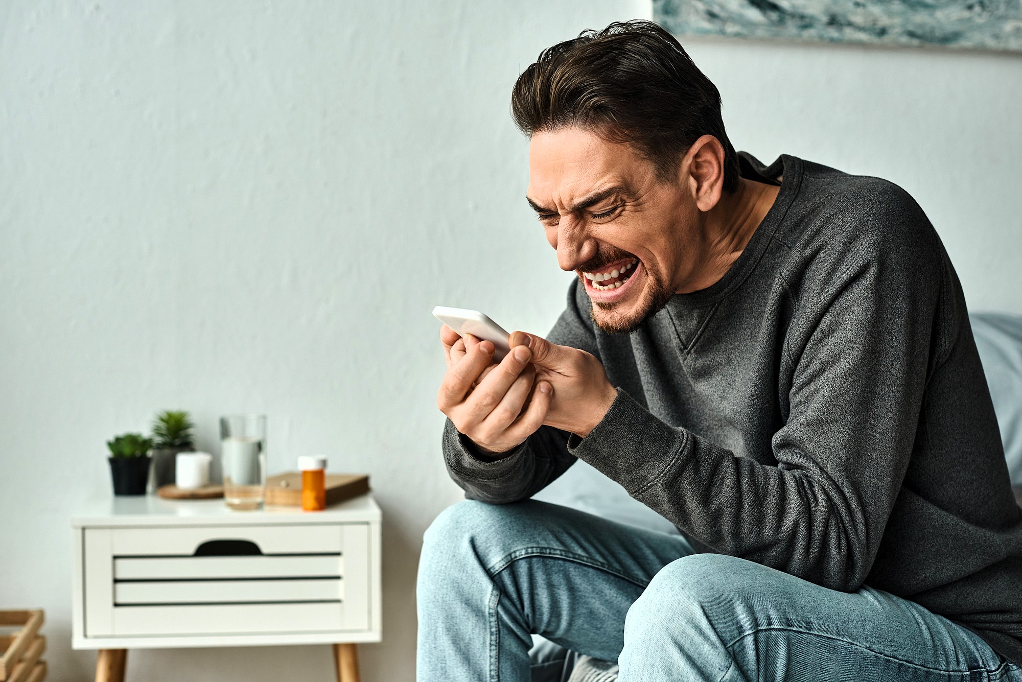 A man in a gray sweater sits on a bed, gripping a smartphone tightly. His expression is intense and frustrated. The bedside table next to him holds various small items, including a plant and a pill bottle.