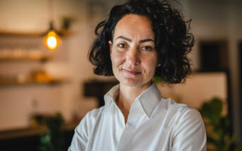 A person with curly dark hair and a white shirt stands indoors, smiling softly. The background is a softly lit room with blurred elements, including a hanging light bulb and shelves.