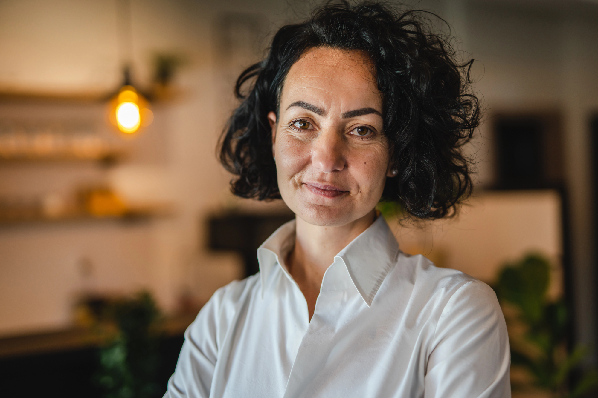 A person with curly dark hair and a white shirt stands indoors, smiling softly. The background is a softly lit room with blurred elements, including a hanging light bulb and shelves.