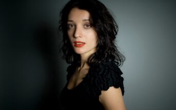 A woman with shoulder-length black hair and red lipstick is posed against a neutral background. She is wearing a black top with ruffled sleeves and looking directly at the camera.