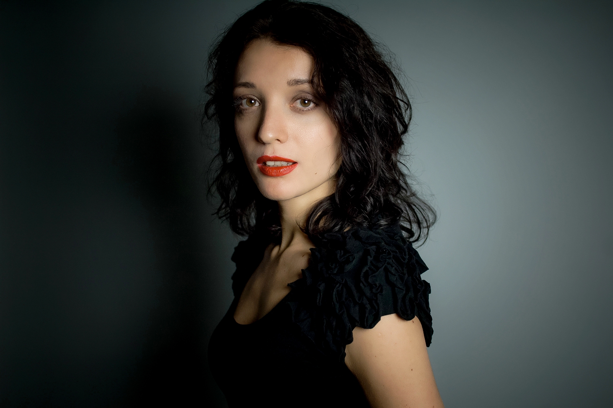 A woman with shoulder-length black hair and red lipstick is posed against a neutral background. She is wearing a black top with ruffled sleeves and looking directly at the camera.