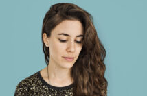 Woman with long, wavy brown hair looks down against a solid light blue background. She is wearing a black top with golden speckles and small stud earrings. Her expression is calm and contemplative.