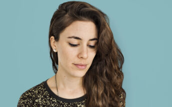 Woman with long, wavy brown hair looks down against a solid light blue background. She is wearing a black top with golden speckles and small stud earrings. Her expression is calm and contemplative.