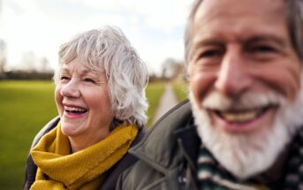 An elderly man and woman are smiling joyfully outdoors. They are wearing warm clothing, with the woman in a yellow scarf. The background features a grassy field and a pathway. The focus is on their happy expressions.
