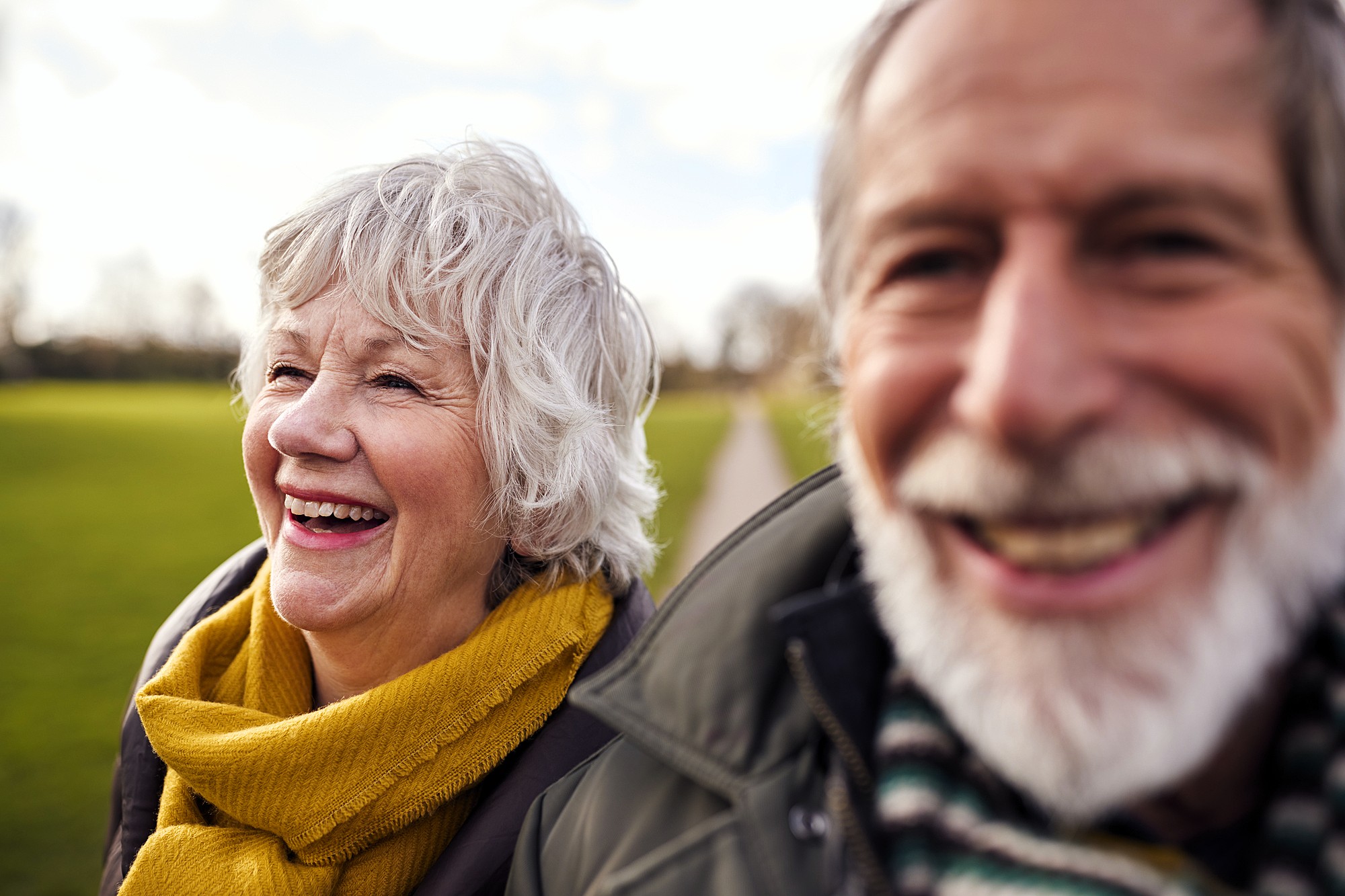 An elderly man and woman are smiling joyfully outdoors. They are wearing warm clothing, with the woman in a yellow scarf. The background features a grassy field and a pathway. The focus is on their happy expressions.