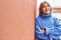 Smiling person with long gray hair wearing a blue turtleneck sweater stands with arms crossed, leaning against a reddish wall. The background is slightly blurred, suggesting an outdoor setting.