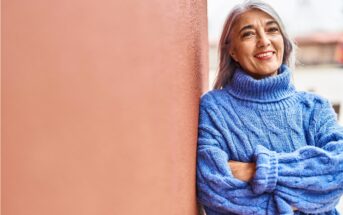 Smiling person with long gray hair wearing a blue turtleneck sweater stands with arms crossed, leaning against a reddish wall. The background is slightly blurred, suggesting an outdoor setting.