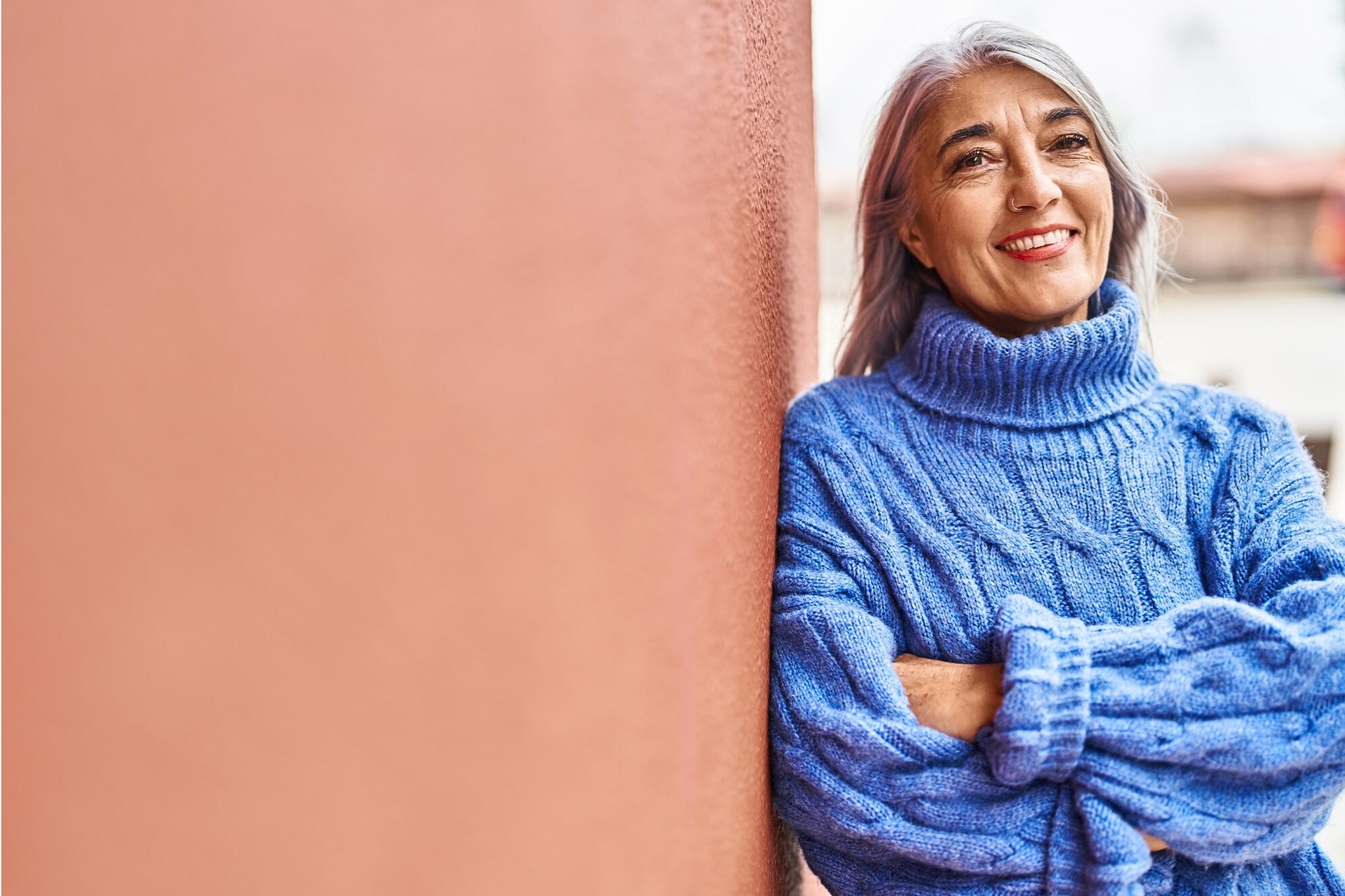 Smiling person with long gray hair wearing a blue turtleneck sweater stands with arms crossed, leaning against a reddish wall. The background is slightly blurred, suggesting an outdoor setting.