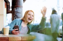 A smiling woman sits at an outdoor café table with a coffee cup and smartphone, enthusiastically waving her hand and greeting someone off-camera.