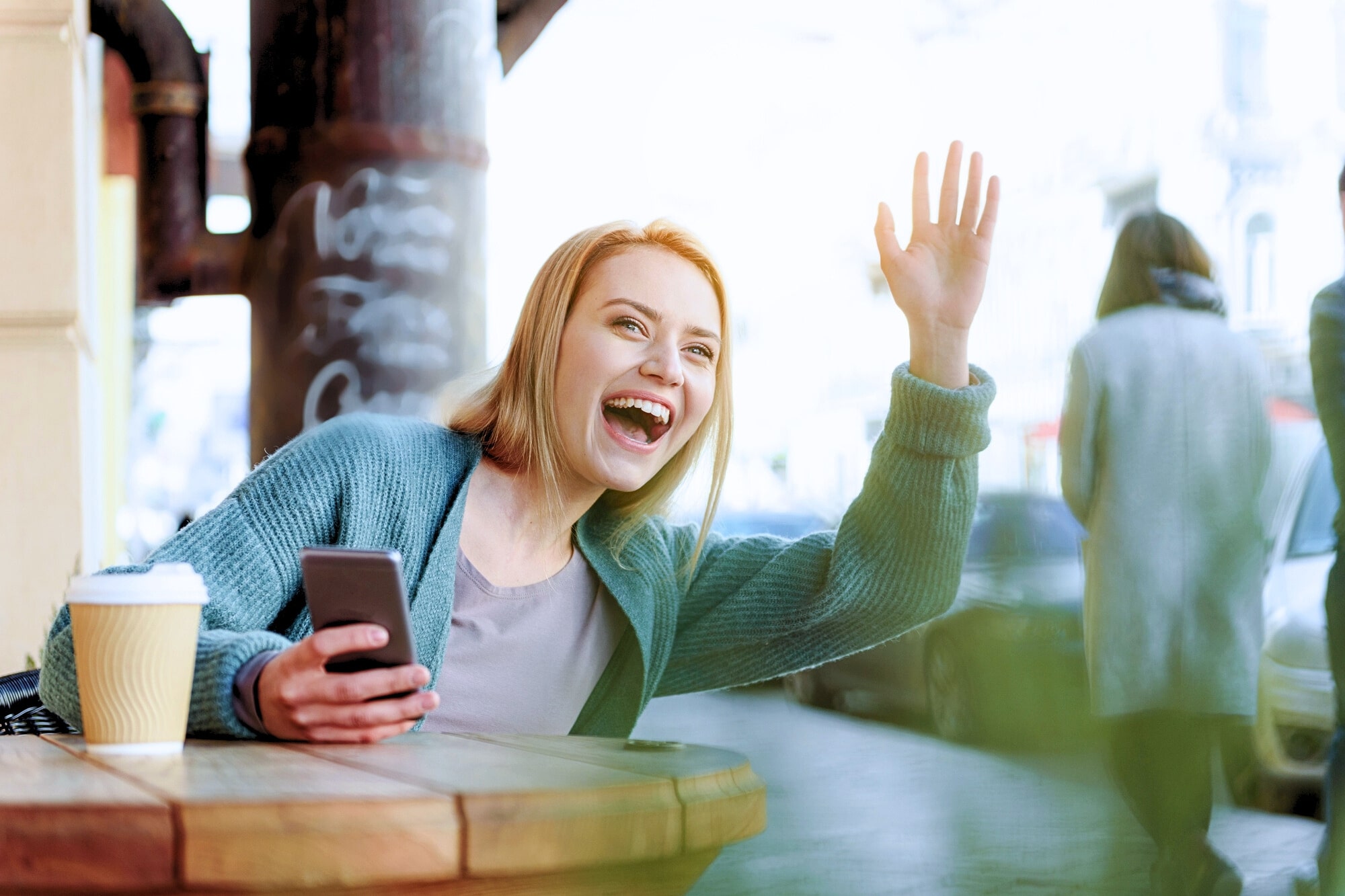 A smiling woman sits at an outdoor café table with a coffee cup and smartphone, enthusiastically waving her hand and greeting someone off-camera.
