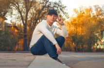 A man in a white sweater, blue jeans, and a black cap sits on a curb in an empty street, surrounded by autumn trees with yellow leaves. He looks at the camera, one hand touching his cap.