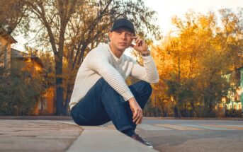A man in a white sweater, blue jeans, and a black cap sits on a curb in an empty street, surrounded by autumn trees with yellow leaves. He looks at the camera, one hand touching his cap.