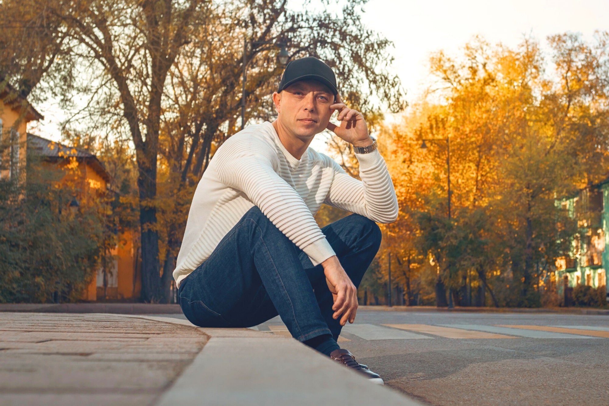 A man in a white sweater, blue jeans, and a black cap sits on a curb in an empty street, surrounded by autumn trees with yellow leaves. He looks at the camera, one hand touching his cap.