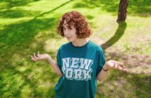 A person with curly hair is standing on a grassy area, wearing a green shirt with "New York" printed on it. They have an expression of uncertainty, with their hands raised and shoulders slightly shrugged. Shadows of trees are visible on the grass.